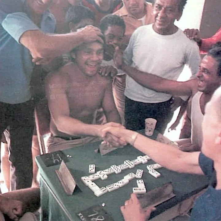 A shirtless man sits at a green domino table, smiling broadly as he shakes hands with an opponent while multiple people…