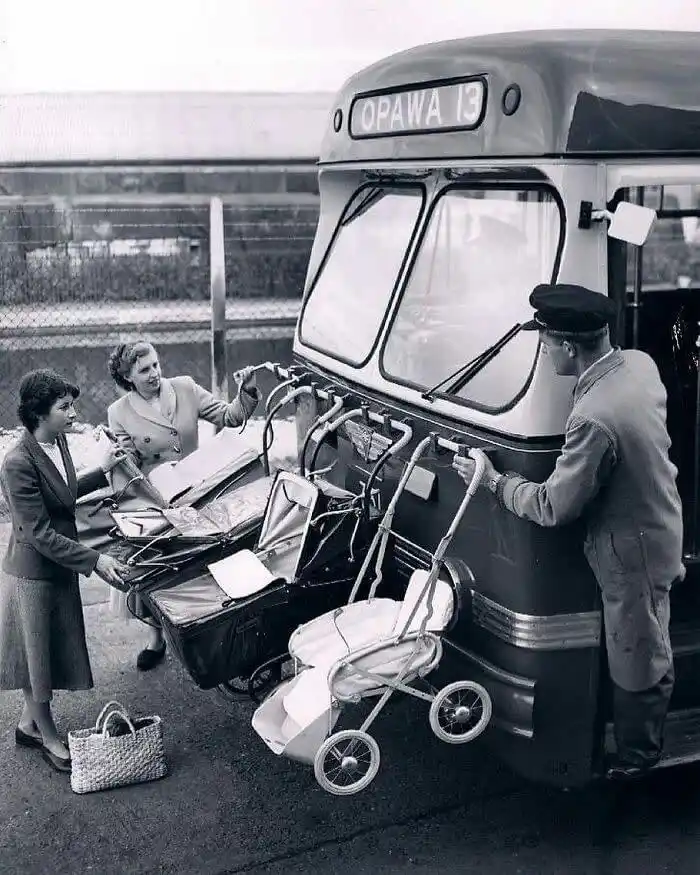 A black-and-white photo shows three women and a man loading multiple baby carriages and strollers onto the front of a…