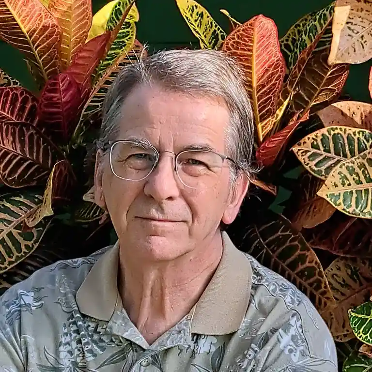 a close-up photo of a middle-aged man with gray hair and glasses, wearing a patterned collared shirt, standing in front of a…