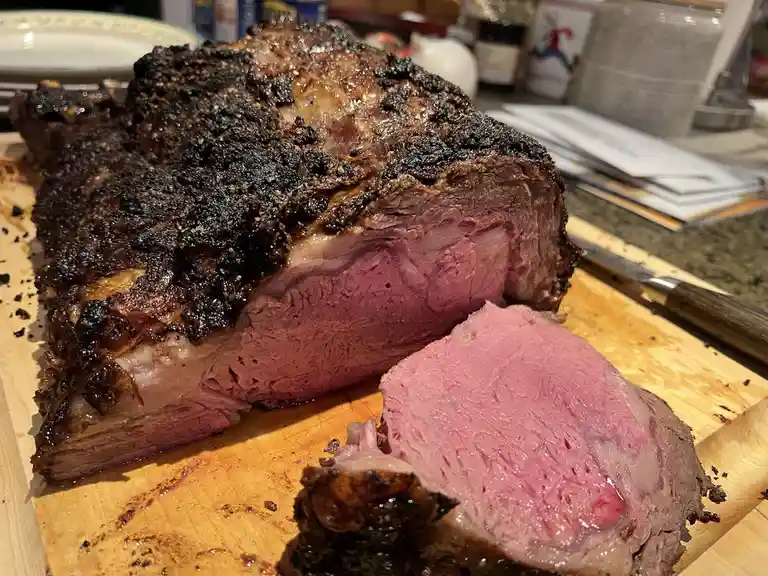 A close-up photo shows a large, sliced roast beef on a wooden cutting board, with a dark, charred exterior and a juicy, pink…