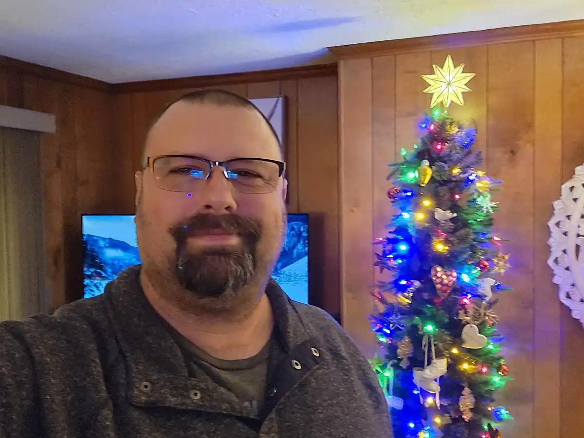 A man with glasses and a goatee takes a selfie in front of a brightly lit, decorated christmas tree with a star on top…