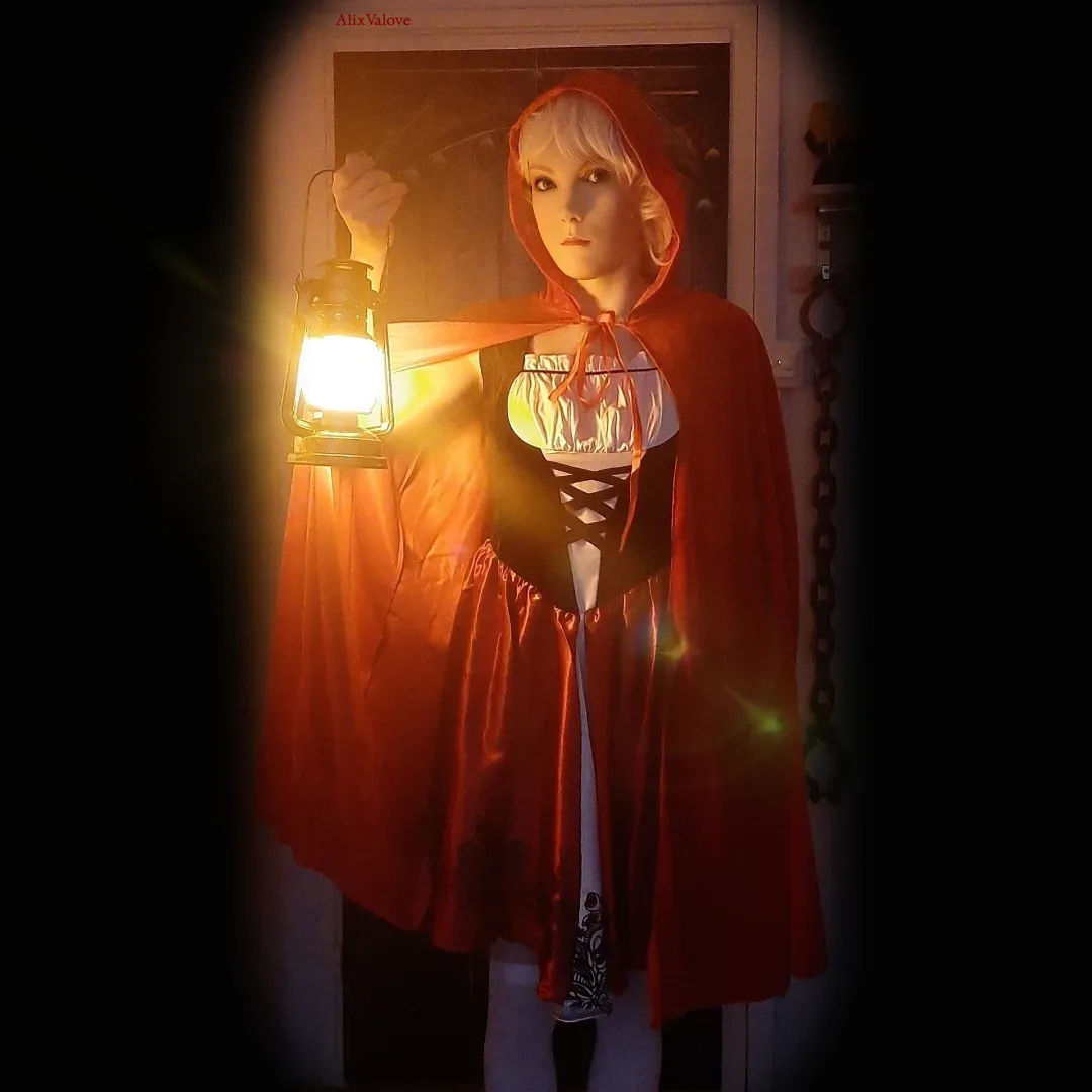 A person with short blonde hair wears a red hooded cape and a black-and-white corset dress, holding a glowing lantern in a…