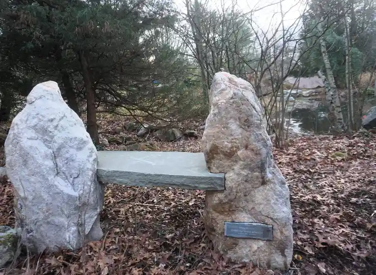 A stone bench with two large, upright rocks as legs sits in a leaf-covered wooded area, with a plaque on the right rock…