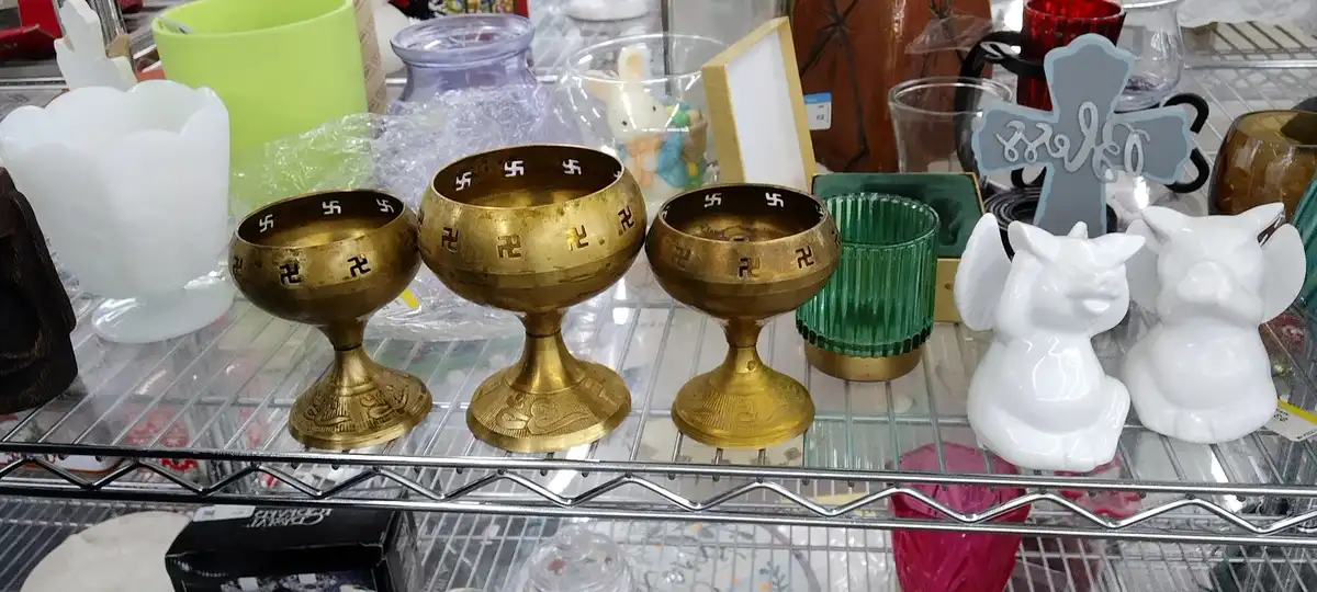 Three golden ceremonial bowls with swastikas engraved around their rims sit on a metal shelf among other decorative items…