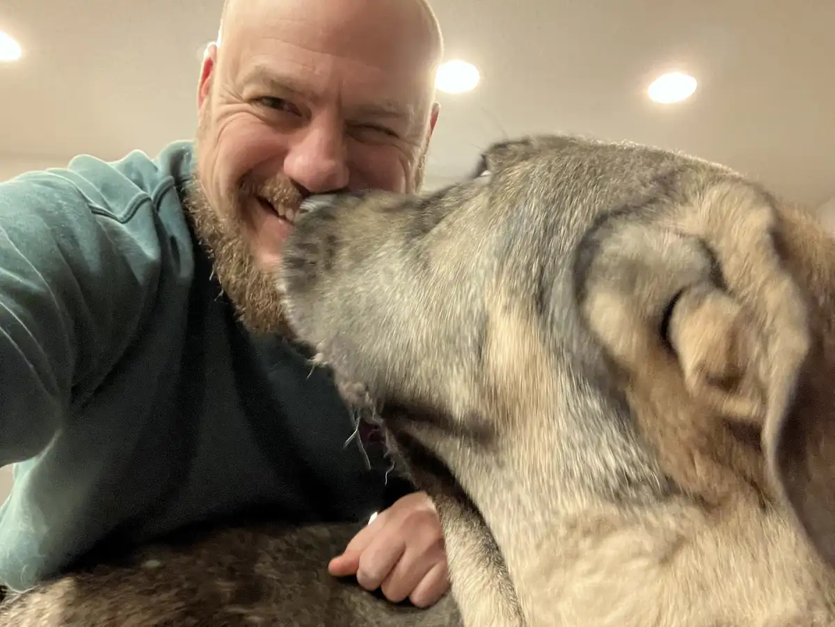 A bald man with a beard in a teal hoodie smiles broadly while taking a selfie, as a large, grey-and-tan dog affectionately…