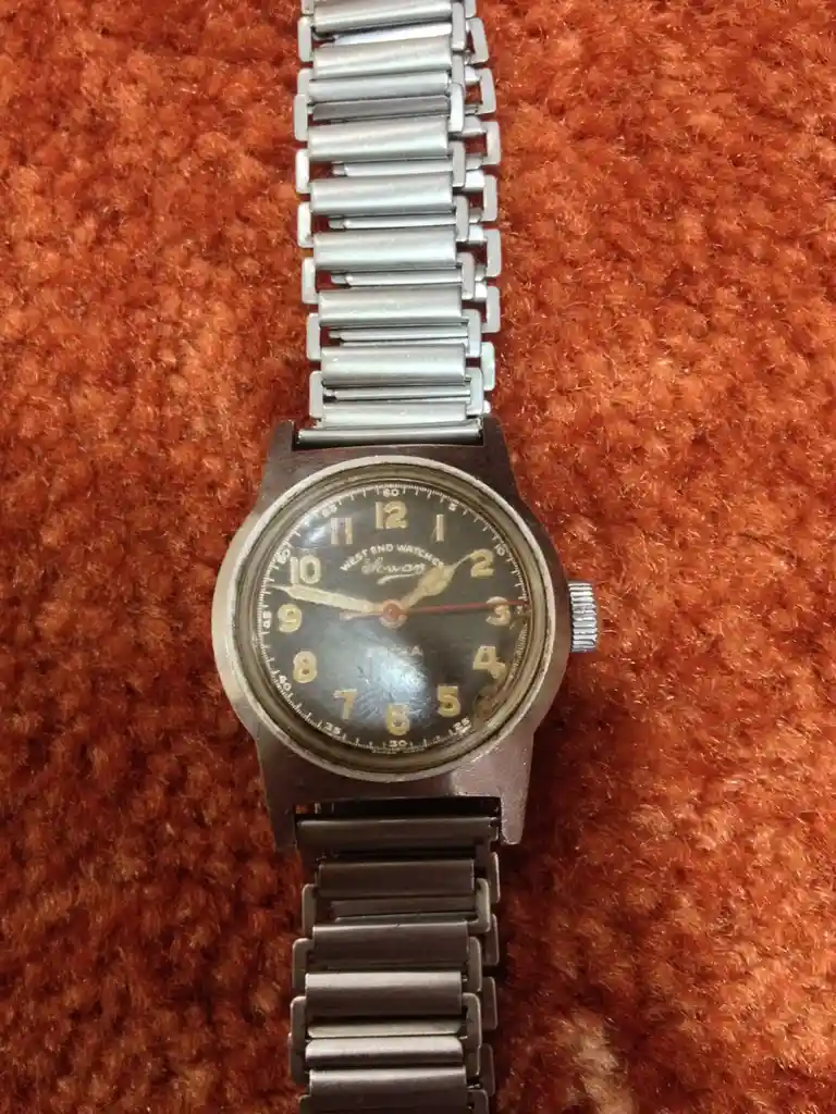 A vintage analog wristwatch with a metal link bracelet rests on a textured orange-brown fabric. The watch face is dark with…