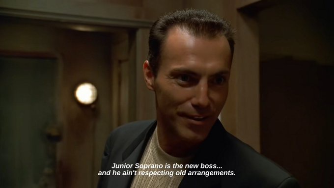 A still from The Sopranos shows a man in a dark suit and light sweater looking intently forward with a serious expression…