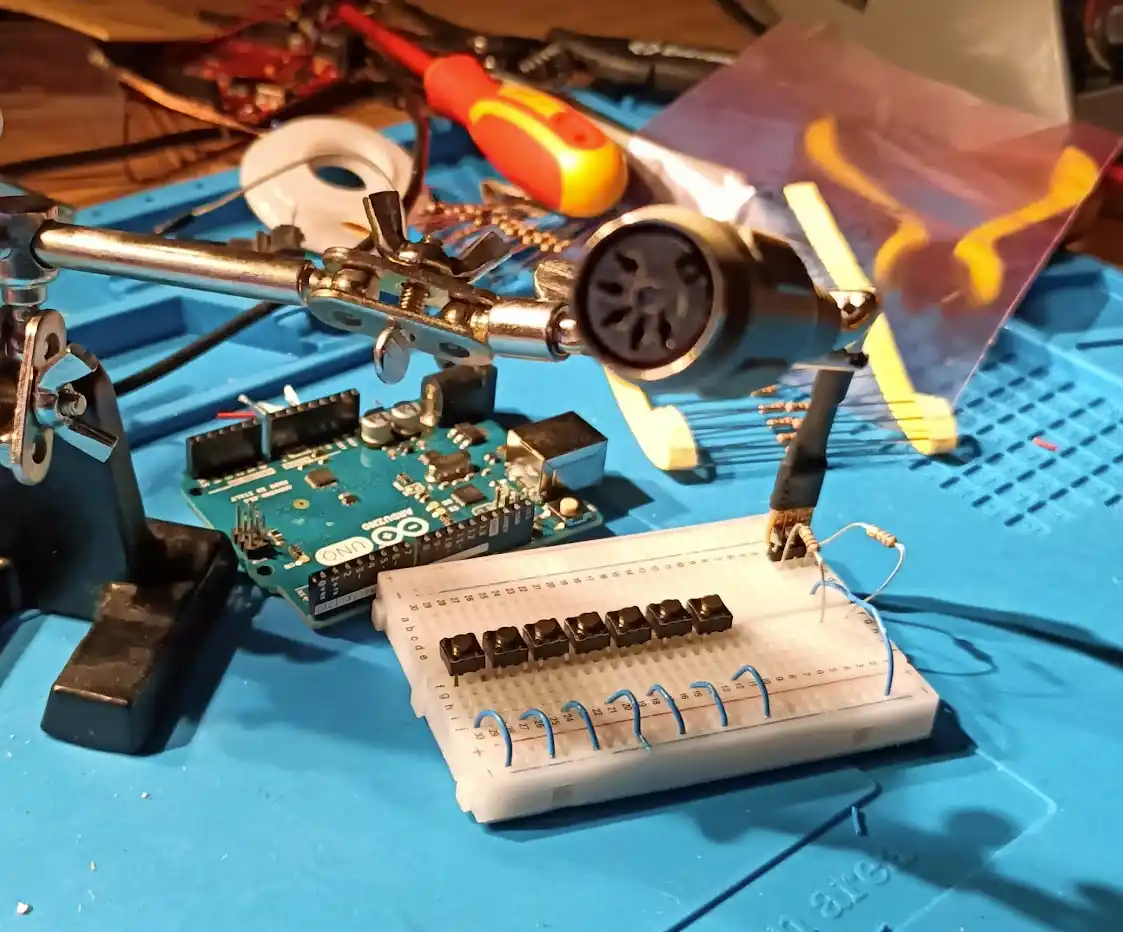 A close-up photo of an electronics workbench with a soldering iron held by a clamp, an Arduino Uno board, a breadboard with…