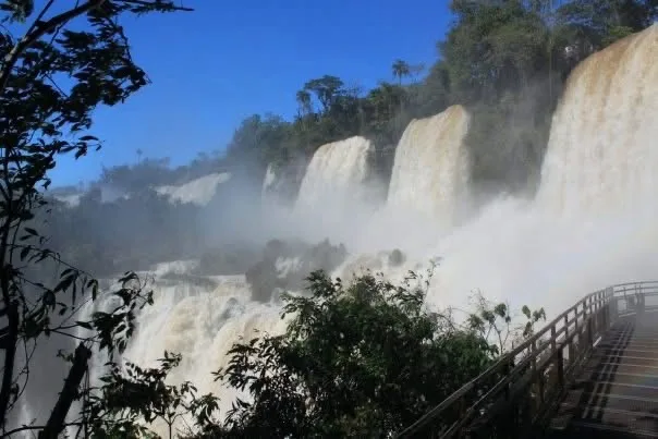 A scenic photo of the Iguazu Falls with mist rising from the cascading water, framed by lush green trees and a wooden…