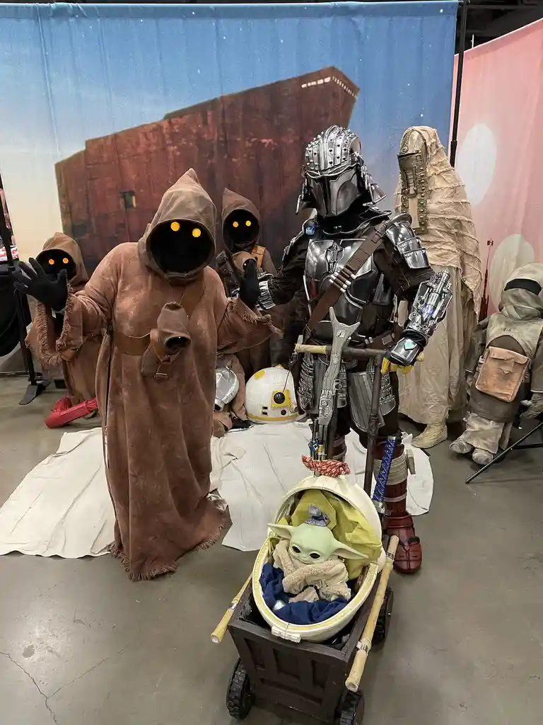 A group of Star Wars cosplayers, including a Mandalorian in full armor pushing a baby carrier with Grogu (Baby Yoda) inside…