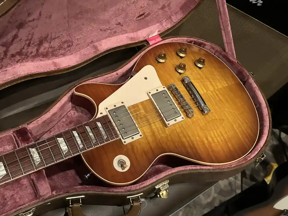 A close-up photo shows a sunburst-finish electric guitar with a white pickguard and gold hardware, resting inside an open…