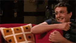 A man in a dark blue shirt sits on a red couch with a patterned pillow, initially smiling and pointing at the viewer, then…