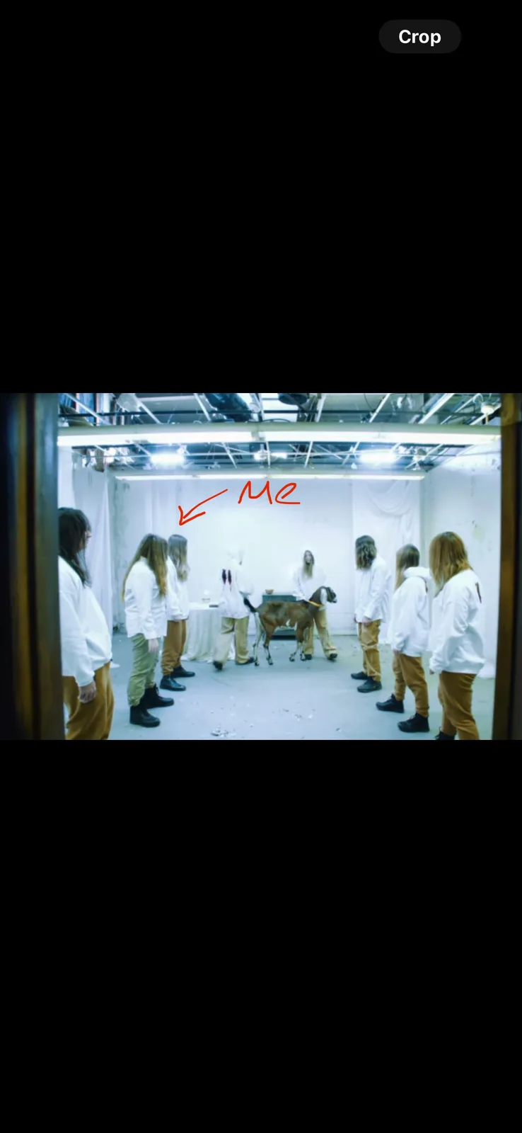 A group of people in white hoodies and tan pants stand in a circle in a brightly lit, white-walled room with exposed ceiling…