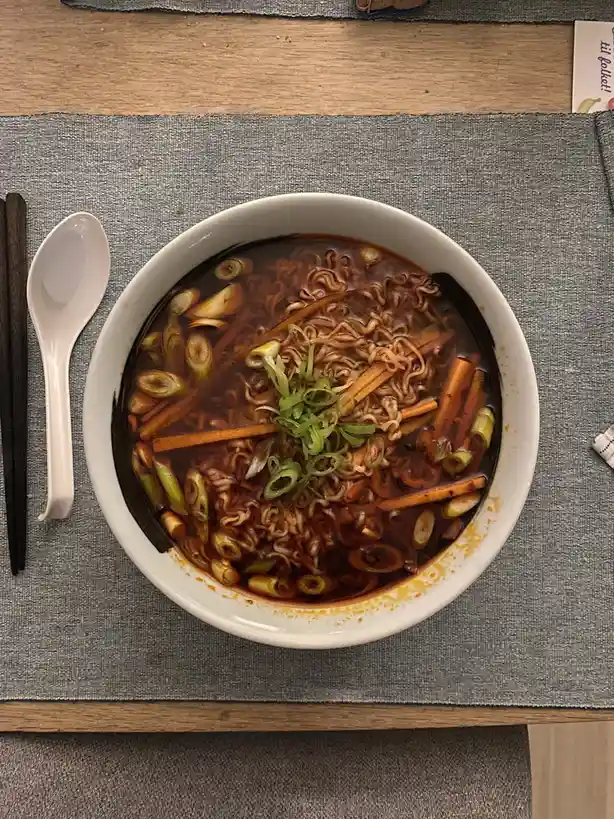 a top-down photo of a white bowl filled with dark broth ramen noodles, garnished with sliced green onions and carrot sticks…