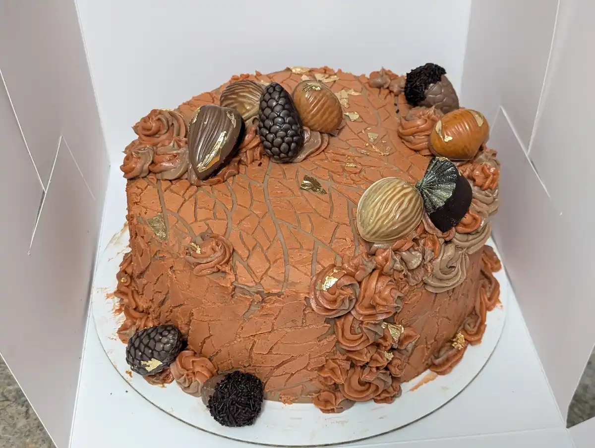 a square chocolate cake with brown frosting, decorated with chocolate-covered nuts, gold leaf, and piped swirls, sits in a…