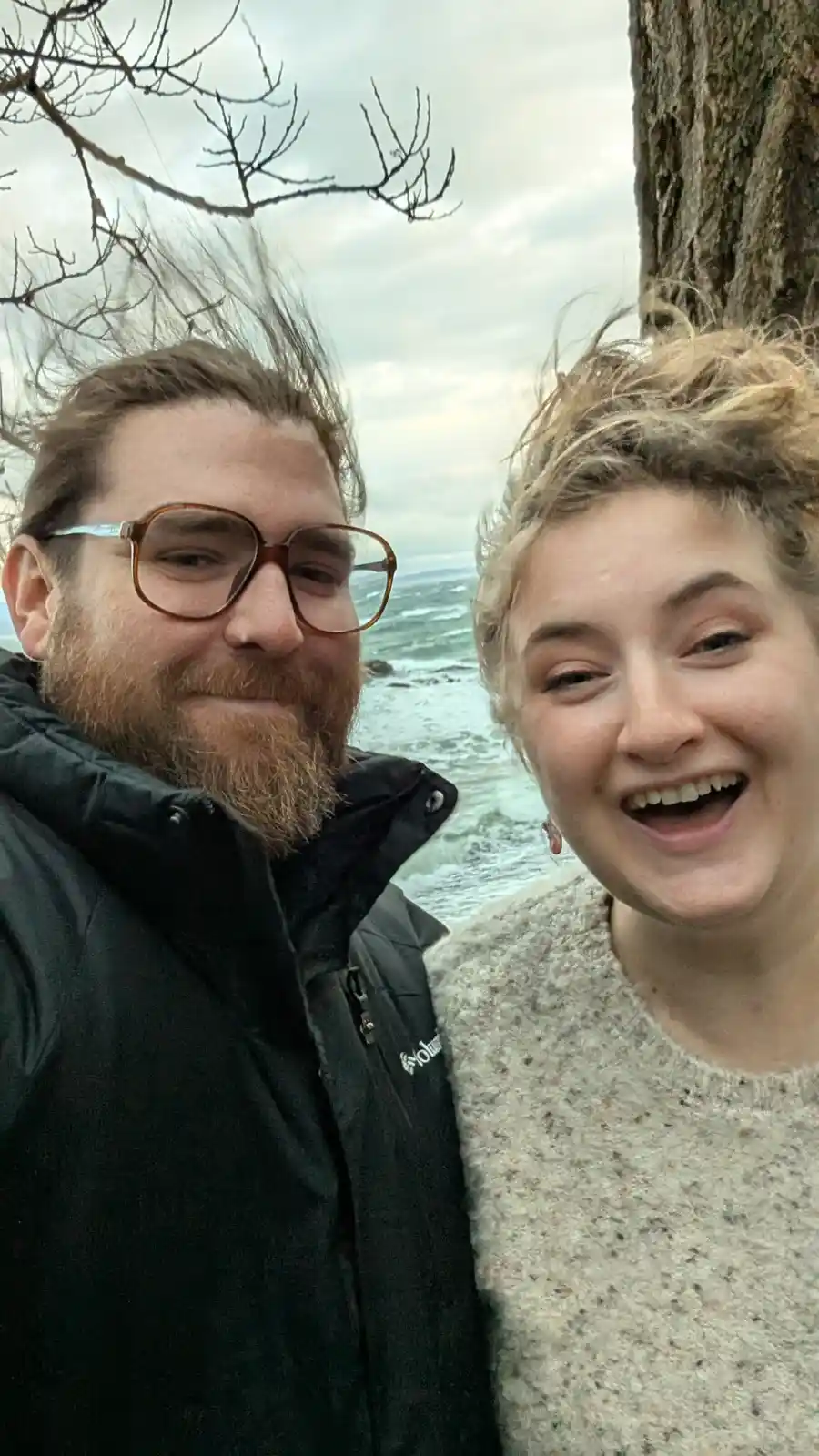 A selfie of a man with glasses and a beard and a woman with blonde hair, both smiling, taken outdoors near a tree with a…