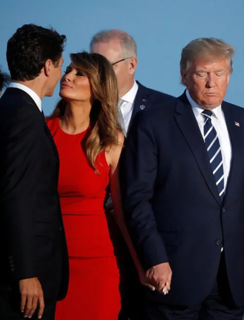 A photo shows Melania Trump in a red dress looking up at a man in a suit while holding hands with Donald Trump, who is…