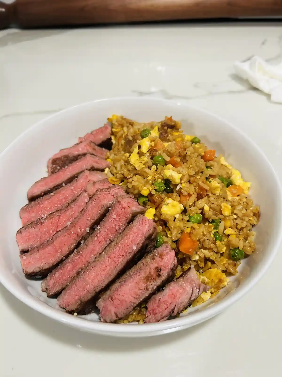 A white bowl contains sliced medium-rare steak seasoned with salt next to fried rice with peas, carrots, and scrambled egg…