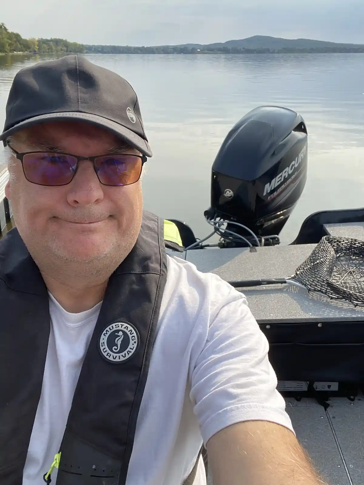 A man wearing a cap, sunglasses, and a Mustang Survival life vest takes a selfie on a boat with a Mercury outboard motor and…