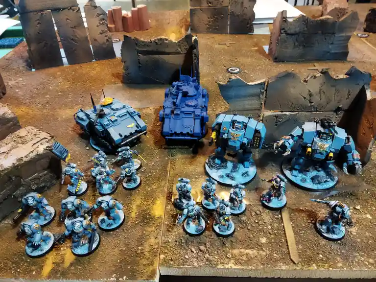 A tabletop wargaming setup featuring numerous blue-painted miniature figures, including tanks and giant robots, arranged on…