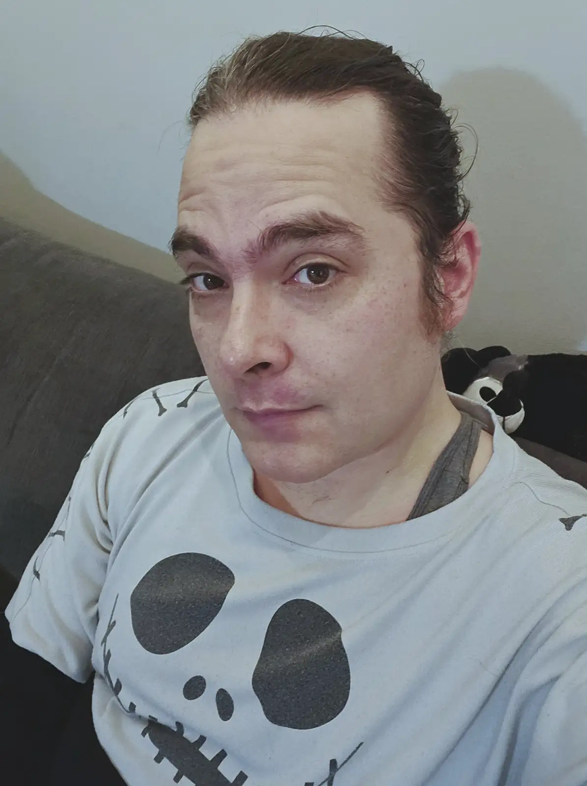 A man with dark hair and freckles sits on a couch wearing a white t-shirt with a large black Jack Skellington face design…