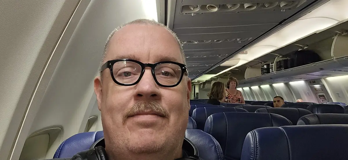 A man with glasses and a mustache takes a selfie inside an airplane cabin, looking directly at the camera with a neutral…