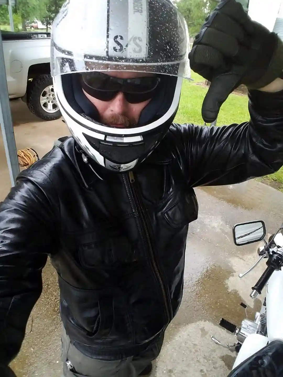 A man in a wet black leather jacket and a full-face motorcycle helmet with a rain-splattered visor gives a thumbs-down…