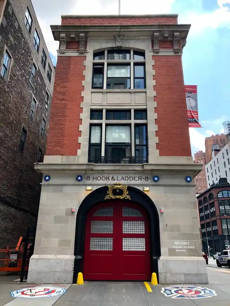 A photo of the exterior of FDNY Engine Company 8, featuring a large red firehouse door under a stone archway with the text…