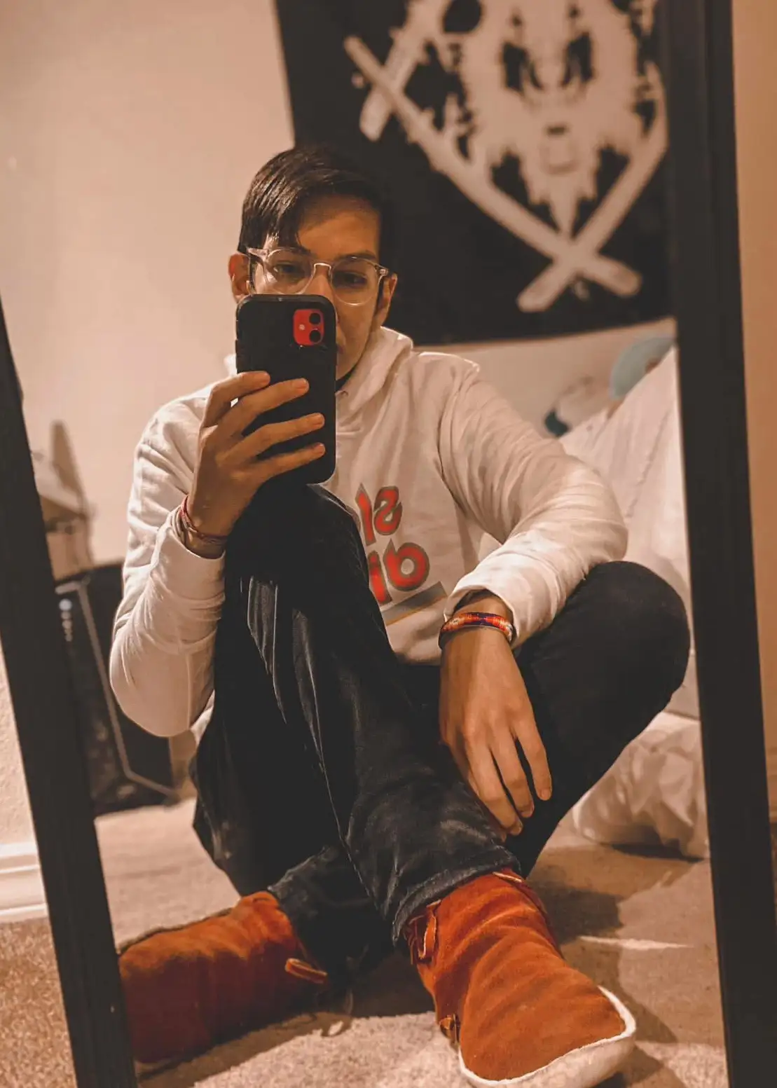 A person with glasses sits cross-legged on a carpeted floor in front of a mirror, holding a black smartphone with a red…