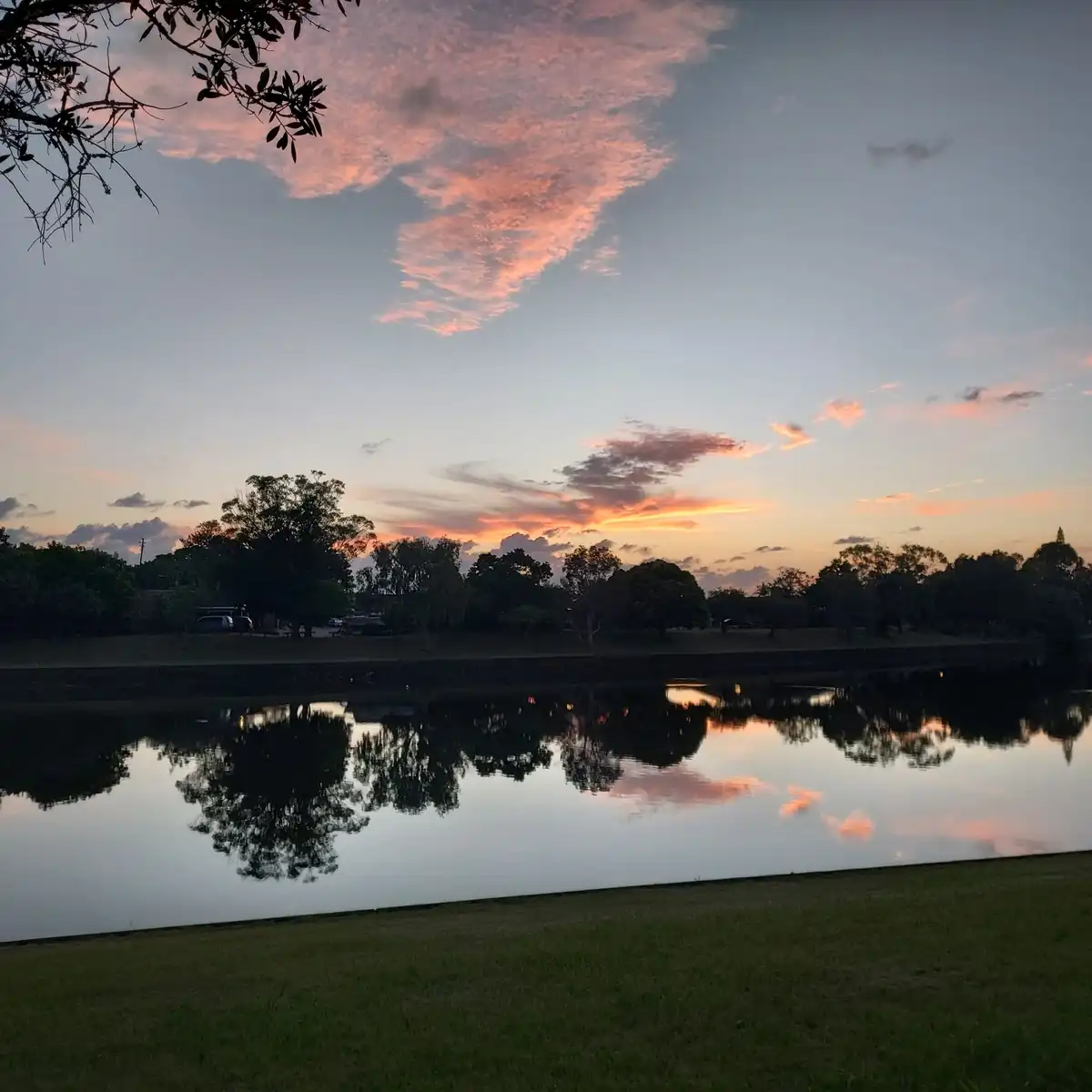 A serene sunset over a calm body of water with trees silhouetted against a pink and orange sky, reflecting in the water. The…