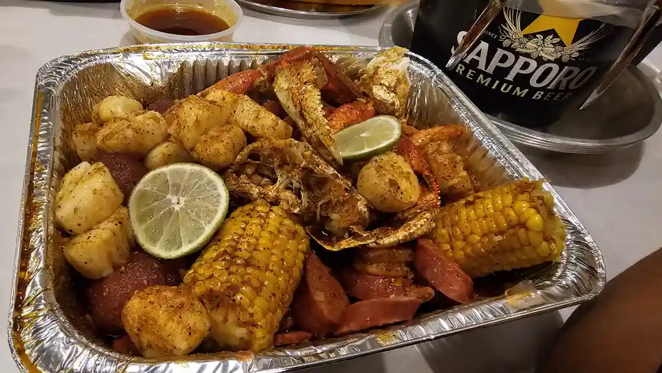 A close-up photo shows a large aluminum foil tray filled with a crawfish boil, containing seasoned crawfish tails, shrimp…
