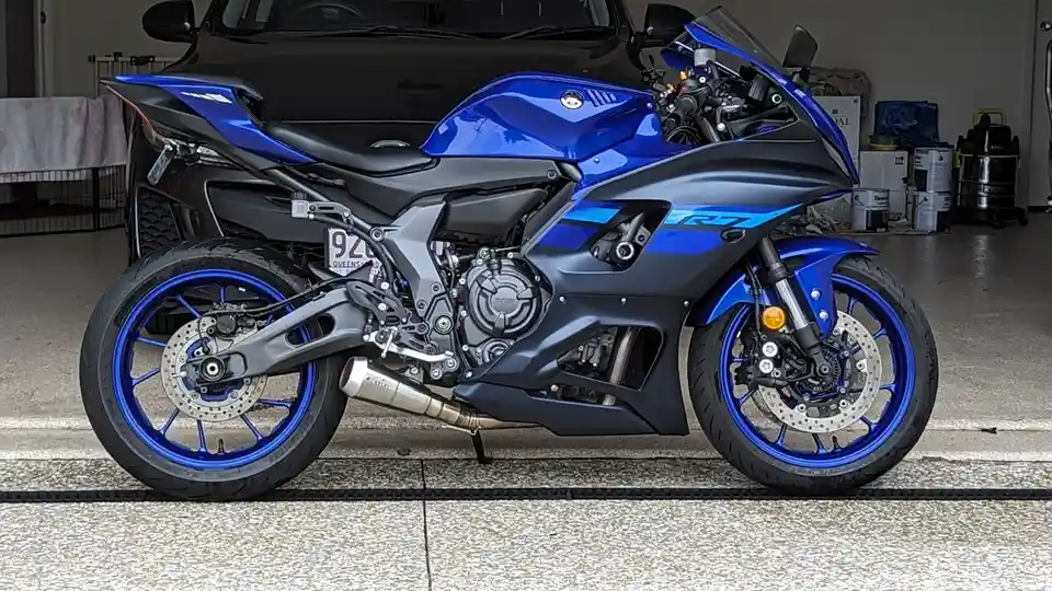 A blue and black Yamaha R7 sport motorcycle is parked in a garage, positioned in front of a black car. The motorcycle has a…