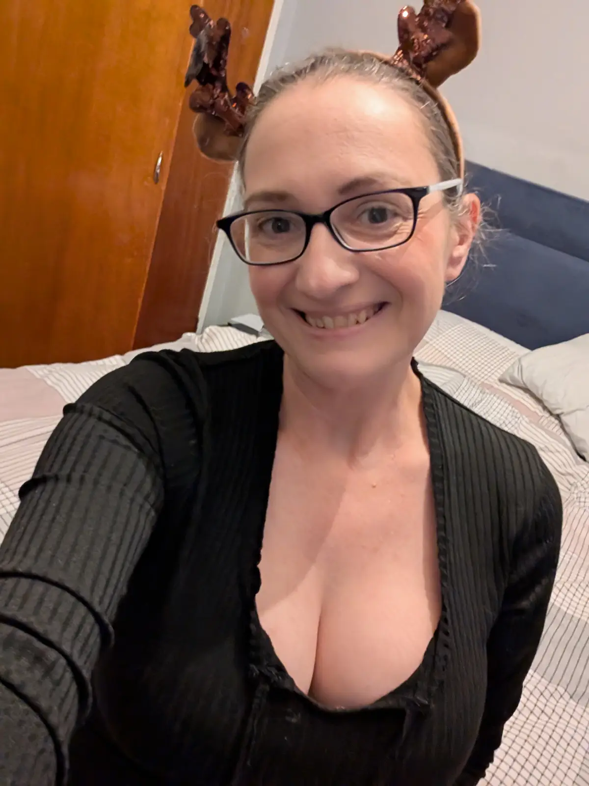 A woman with glasses and a black ribbed top sits on a bed with a striped sheet and blue headboard, wearing shiny brown…