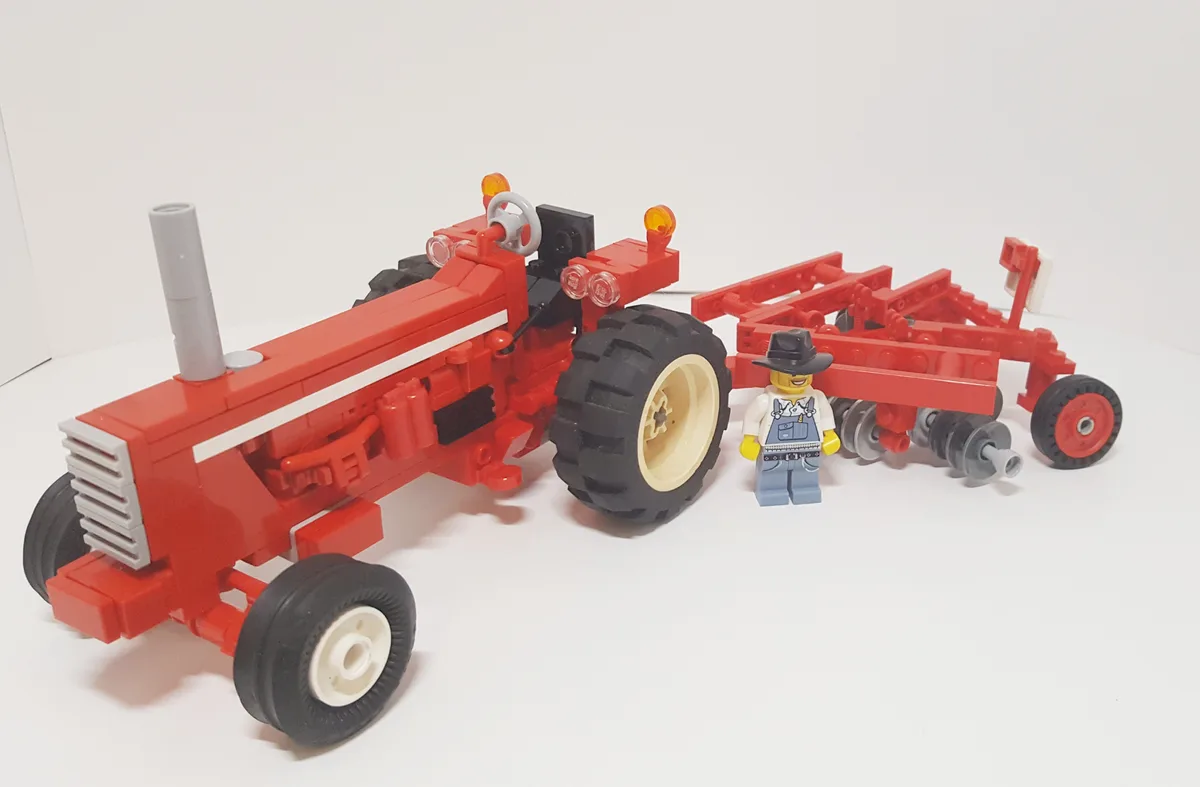 A red Lego tractor with black tires and a gray exhaust pipe is shown next to a red Lego plow, with a Lego minifigure wearing…