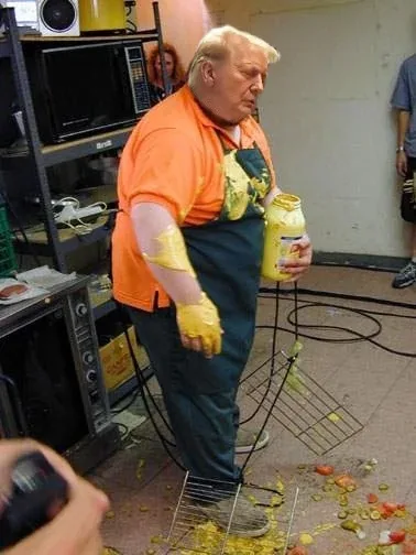 A man with distinctive blonde hair and an orange shirt is covered in yellow mustard, holding a jar of mustard, with mustard…