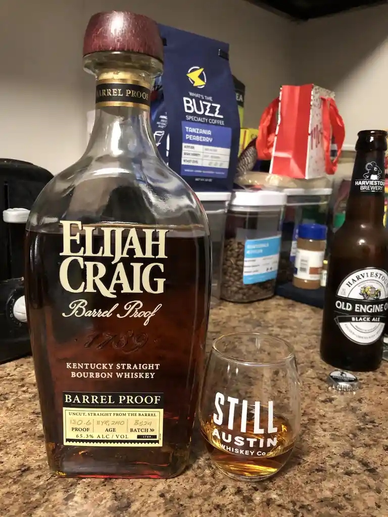 A bottle of Elijah Craig Barrel Proof bourbon whiskey sits next to a glass labeled 'STILL AUSTIN WHISKEY CO' containing…