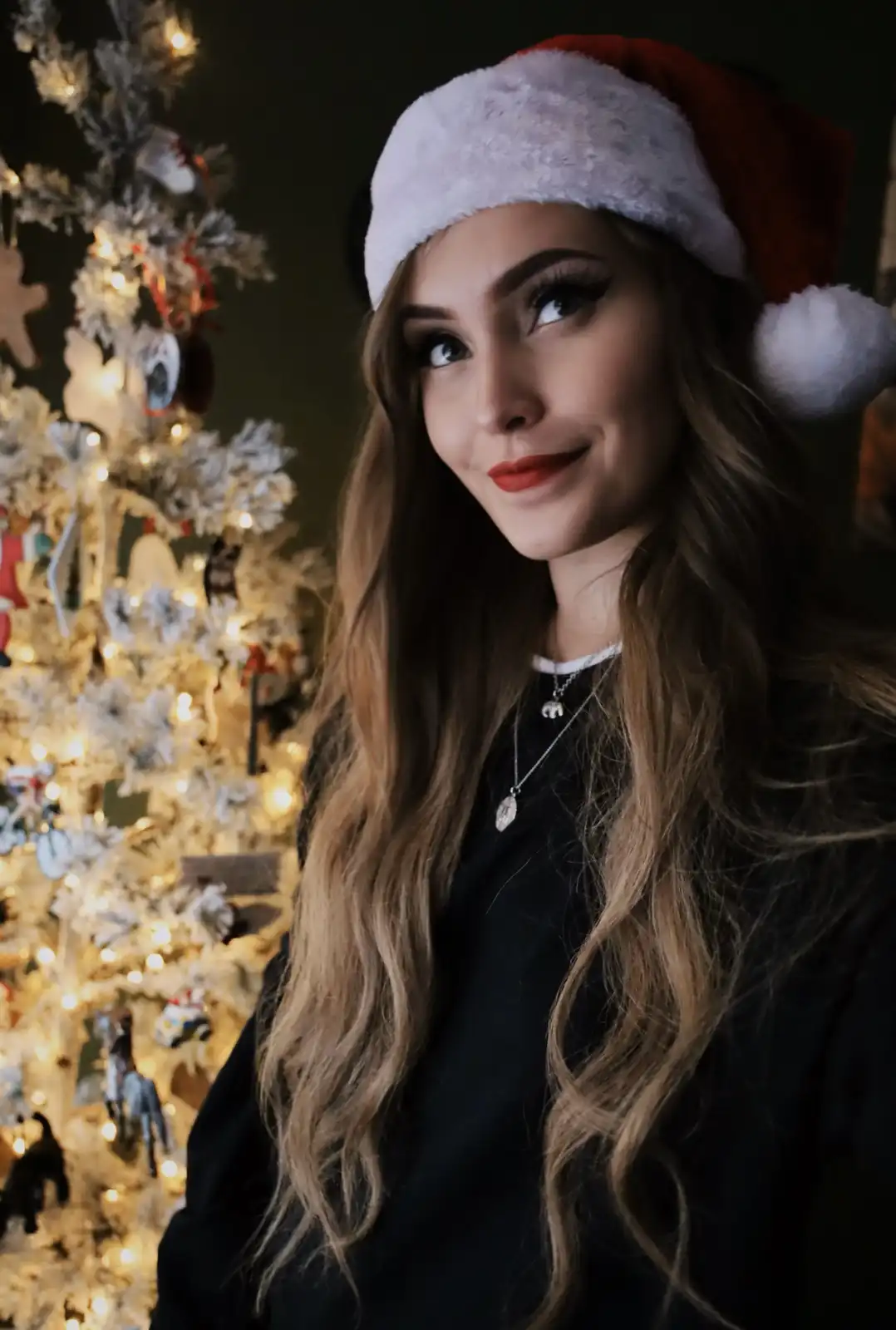 A woman with long blonde hair wearing a red and white Santa hat and a black top stands in front of a decorated, lit…