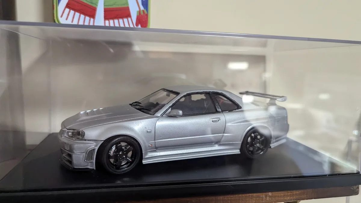 A silver die-cast model of a Nissan Skyline GT-R R34 is displayed inside a clear plastic case on a black base. The car has…
