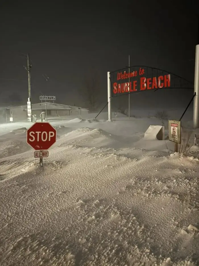A nighttime photo of a snow-covered town entrance with a large red 'Welcome to Sauble Beach' sign and a stop sign partially…