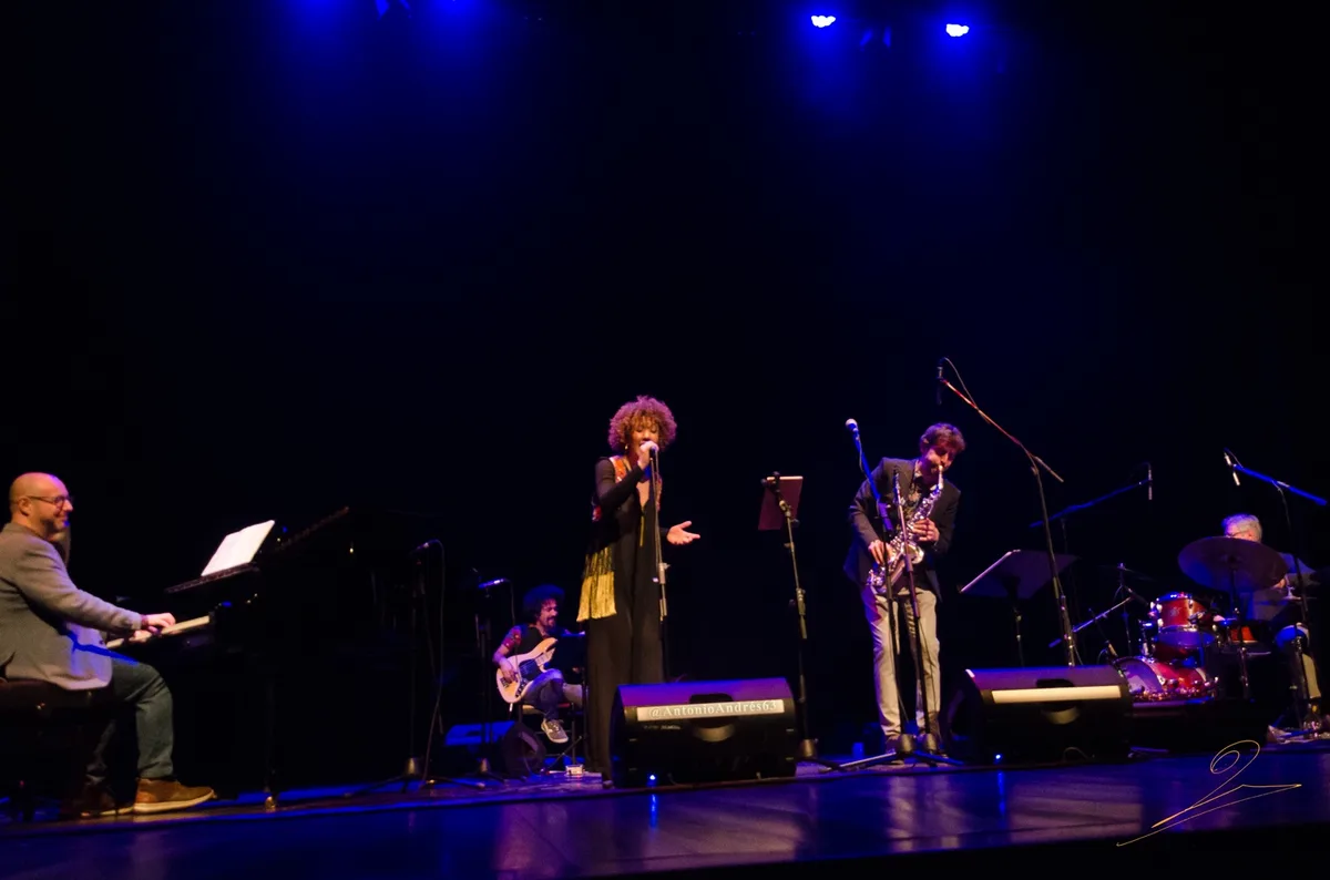 A live jazz band performs on a dimly lit stage under blue spotlights, featuring a singer, saxophonist, pianist, bassist, and…