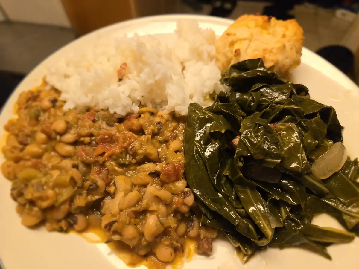 A close-up photo of a plate of food featuring black-eyed peas, collard greens, white rice, and a biscuit. The overlaid text…