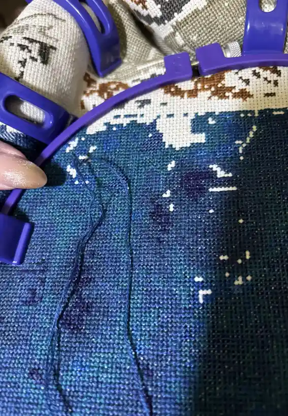 A close-up photo shows a hand holding a purple embroidery hoop with a partially completed cross-stitch project. The fabric…