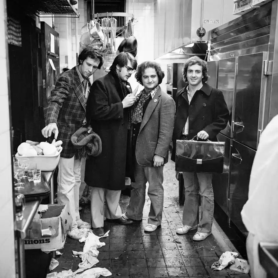 A black-and-white photo shows four men in 1970s-style clothing standing in a messy, cluttered commercial kitchen or food…
