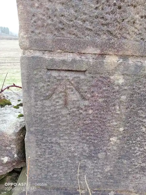A close-up of a weathered, grey stone monument outdoors, with faint, eroded carvings visible on its surface. The overlaid…