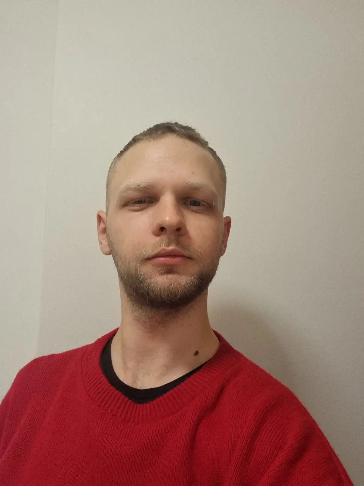 A man with short hair and a beard, wearing a red sweater with a black collar, stares blankly at the camera against a plain…