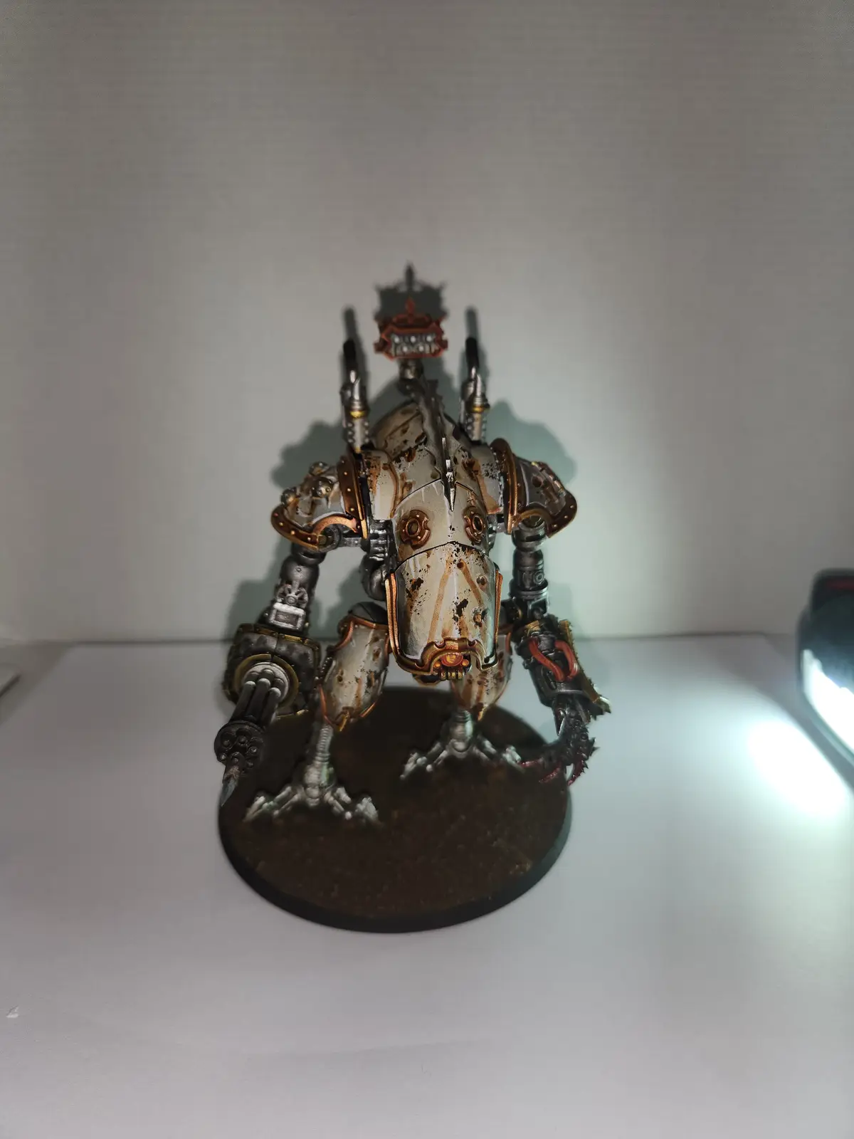 A heavily weathered, custom-painted miniature robot figurine with rust and battle damage stands on a circular base against a…