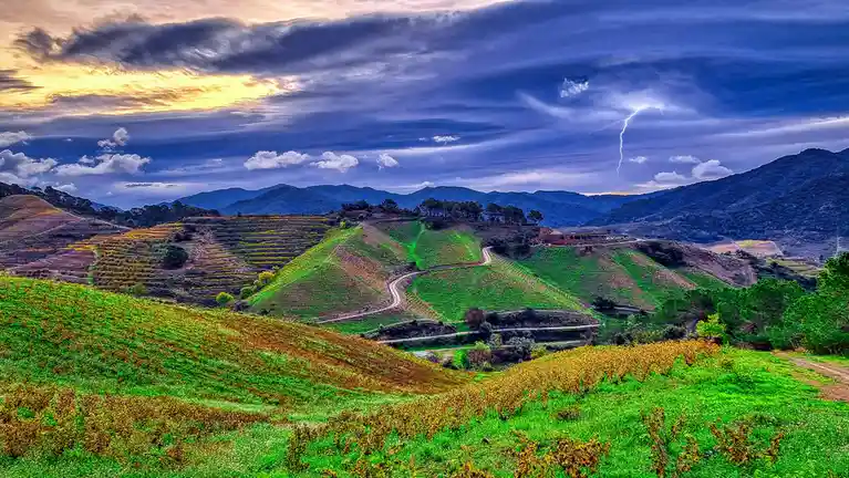 A vibrant, heavily saturated landscape photo shows rolling hills with terraced vineyards under a dramatic sky. A bright…