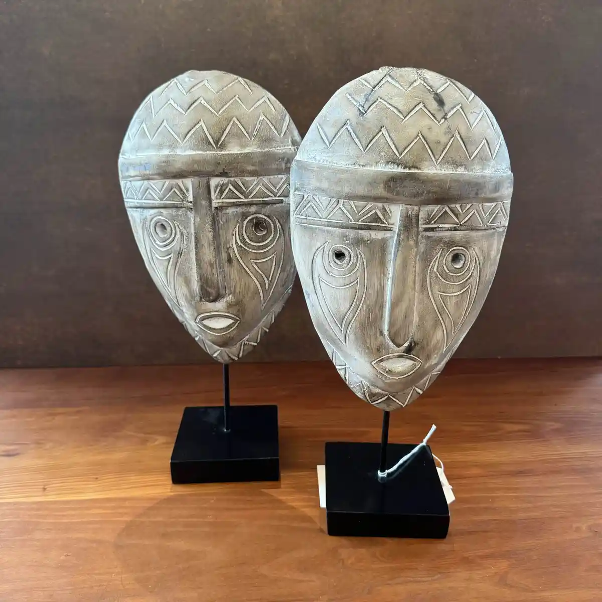 Two identical wooden tribal-style masks with zigzag patterns and stylized eyes are displayed on black stands on a wooden…