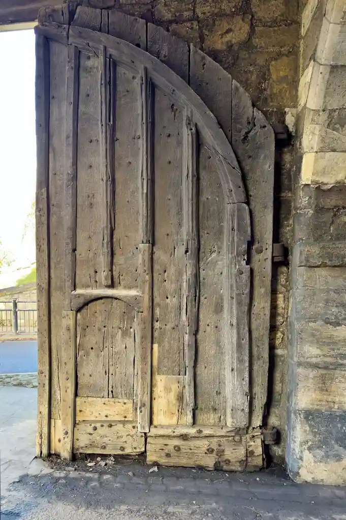 A weathered, ancient wooden door with a curved top, showing significant wear, cracks, and nail holes, set in a stone…