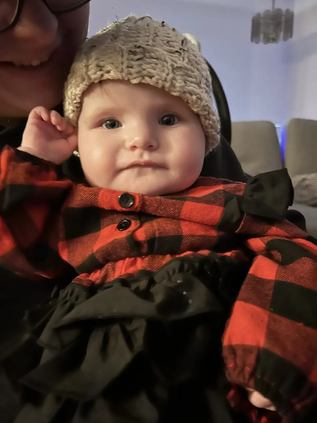A close-up photo shows a baby wearing a beige knit hat and a red-and-black plaid outfit with black bows, looking directly at…